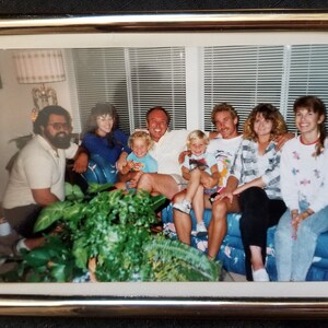 May include: A group of seven people are sitting on a couch. There are three adults and four children. The adults are all smiling and the children are looking at the camera. There are plants in the foreground.