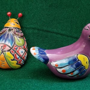 May include: A ceramic ladybug figurine with a yellow, blue, and red patterned shell and two red antennae, positioned next to a purple ceramic bird figurine featuring a blue and yellow wing design with orange accents and closed eyes.