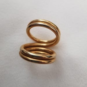 May include: A gold-toned ring featuring a continuous, spiraling design that wraps around itself to form two distinct circular loops. The metal surface is smooth and polished, reflecting light across the curved, rounded edges of the band.