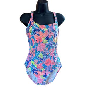 May include: A one-piece swimsuit featuring a vibrant, multi-colored tropical floral and botanical print. The design includes shades of bright pink, neon orange, lime green, and deep purple set against a vivid blue background with a scoop neckline.