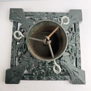 May include: A square dark green cast iron Christmas tree stand featuring an ornate openwork pattern. The central cylindrical holder includes three adjustable metal eye bolts with threaded shafts to secure a tree trunk in place.