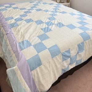 May include: A patchwork quilt featuring a repeating geometric pattern of light blue and off-white squares. The quilt includes a border of solid lavender fabric along one side and is draped over a bed, showcasing a stitched grid construction.