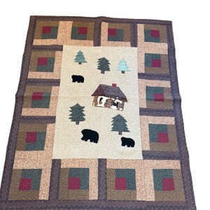 May include: A rectangular quilt featuring a central cream-colored panel with appliquéd black bear silhouettes, evergreen trees, and a small cabin. The border consists of a repeating log cabin block pattern in shades of burgundy, tan, and dark green.