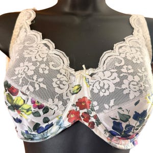 May include: A white underwire bra featuring a floral pattern with red, blue, yellow, and green flowers on the lower cups. The upper cups are adorned with white lace trim in a floral motif, and a small white bow is centered at the bridge.