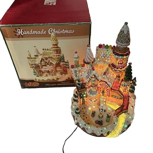 May include: A light-up gingerbread house figurine with a central carousel, multi-colored LED lights, and candy-themed architectural details. It is accompanied by a red and white cardboard box labeled Handmade Christmas with a picture of the house.