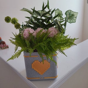 May include: A grey planter with a yellow pixelated heart design, filled with green ferns, pink flowers, and small green plants.