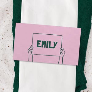 Puede incluir: Un lugar en la mesa con un mantel individual tejido, un plato moteado y una servilleta blanca doblada. Una tarjeta de nombre rosa con el nombre "EMILY" en letras verdes es sostenida por manos ilustradas, descansando sobre una servilleta verde.