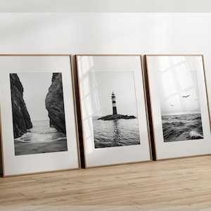 May include: Three framed black and white landscape prints. The prints depict a rocky coastline, a lighthouse on a small island, and a seascape with birds. The frames are a light brown color, and the images are displayed on a wooden floor.