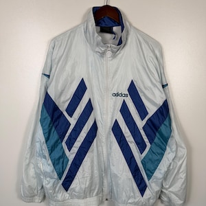 May include: A vintage Adidas track jacket in light blue with navy and teal geometric accents. The jacket features a full zipper, a high collar, and the Adidas logo on the chest. The jacket is hanging on a wooden hanger.