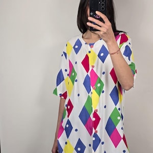 May include: A white button-down shirt with a colorful harlequin pattern of diamonds in pink, blue, green, and yellow. The shirt has short sleeves and pink buttons. The shirt is a tunic style.