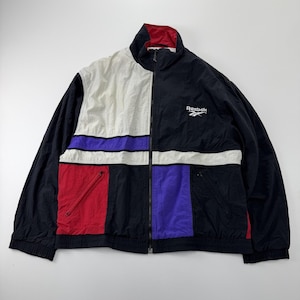 Può includere: Una giacca a vento Reebok caratterizzata da un design a blocchi di colore nero, bianco, rosso e viola. La giacca presenta un colletto alto, una chiusura frontale con zip, maniche lunghe con polsini elastici e due tasche con zip sul davanti.