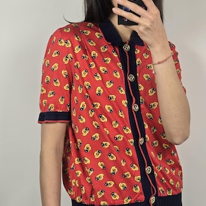 May include: Red short-sleeved button-up shirt with a repeating yellow and black pattern. The shirt has navy blue trim on the collar, sleeves, and button placket. Gold-colored buttons are visible.