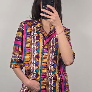 May include: A colorful button-down shirt with a vibrant geometric pattern. The shirt features vertical stripes in shades of pink, yellow, teal, and purple. The short-sleeved shirt has a collared neckline and a pocket on the chest.