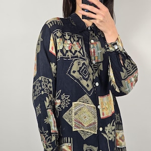 May include: Black button-down shirt with a colorful geometric pattern. The long-sleeved shirt features a collared neckline and white buttons. The pattern includes squares, rectangles, and floral designs in shades of green, red, and gold.