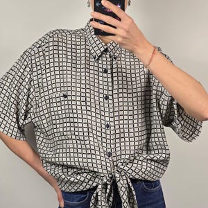 May include: A short-sleeved button-down shirt with a black and white geometric pattern, tied at the waist. The shirt has a small pocket on the chest and is paired with blue jeans.