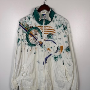 May include: White zip-up jacket with a green collar and abstract design in green, blue, and orange. The jacket has two front pockets and a ribbed hem and cuffs. The jacket is hanging on a wooden hanger.