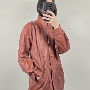 May include: A reddish-brown leather jacket with a high collar and button closure. The jacket features long sleeves, pockets, and a tailored fit. The vintage jacket is a stylish outerwear piece.