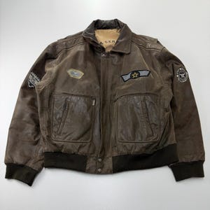 May include: A brown leather bomber jacket featuring a tan interior lining, dark brown ribbed cuffs and hem, and multiple embroidered patches on the chest and sleeves. The jacket includes two large front pockets with zippers and a central snap closure.