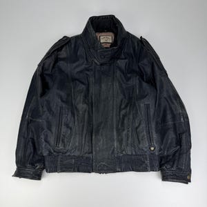 Puede incluir: Una chaqueta de cuero azul marino oscuro con cuello alto, charreteras en los hombros y bolsillos verticales con cremallera. La etiqueta interior muestra el texto Camel. La chaqueta incluye dobladillo y puños acanalados, con cierre frontal.