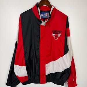 May include: A Chicago Bulls windbreaker jacket in red, black, and white. The jacket features the team logo on the chest and a Starter logo on the collar. The jacket is hanging on a wooden hanger.