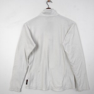 Op de afbeelding: Een witte fleecejas met een hoge kraag en lange mouwen hangt aan een houten kleerhanger. De jas heeft een rits die tot aan de kraag gaat. De jas is gemaakt van een zacht, warm materiaal.