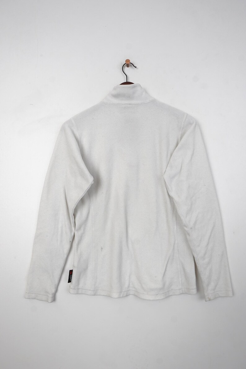 Op de afbeelding: Een witte fleecejas met een hoge kraag en lange mouwen hangt aan een houten kleerhanger. De jas heeft een rits die tot aan de kraag gaat. De jas is gemaakt van een zacht, warm materiaal.