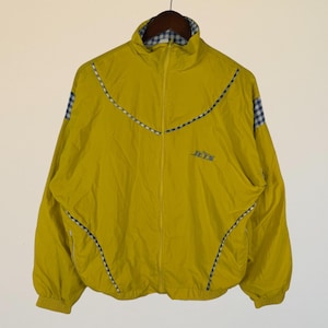 May include: Mustard yellow vintage jacket with a full zipper and a stand-up collar. The jacket features blue and white checkered accents on the shoulders and collar, with the word "JETS" printed on the front.