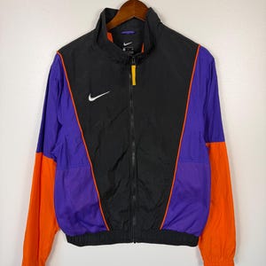 May include: Black, purple, and orange athletic jacket with a full-length zipper and a white Nike logo on the chest. The jacket has a high collar and orange mesh detailing on the sides. The sleeves are orange, and the jacket is hanging on a wooden hanger.