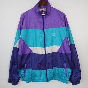 May include: A vintage windbreaker jacket in a color block design. The jacket features a high collar, a full-length zipper, and a logo on the chest. The color scheme includes purple, teal, white, and navy blue. The jacket is hanging on a wooden hanger.