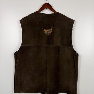 May include: A brown suede vest featuring an embroidered pheasant design on the back. The sleeveless vest is suspended from a wooden hanger. The pheasant embroidery uses brown, green, and gold thread against the brown suede.