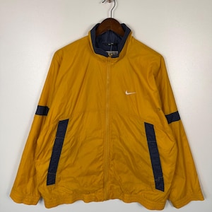 May include: Mustard yellow Nike track jacket with navy blue accents. Features a full zipper, a small white Nike logo on the chest, and navy blue stripes on the sleeves. The jacket has a navy blue collar and pocket details.