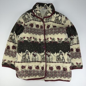 Könnte beinhalten: Eine cremefarbene Fleecejacke mit einem sich wiederholenden Muster aus Teddybären, Pinguinen und dunkelroten Rosen. Die Jacke hat eine Knopfleiste vorne, einen Stehkragen und dunkelrote Einfassungen an den Rändern und Ärmelbündchen.