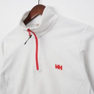 Op de afbeelding: Witte fleece trui met een hoge kraag en een rode rits. De trui heeft de geborduurde rode letters "HH" op de borst. Het kledingstuk hangt aan een houten kleerhanger.