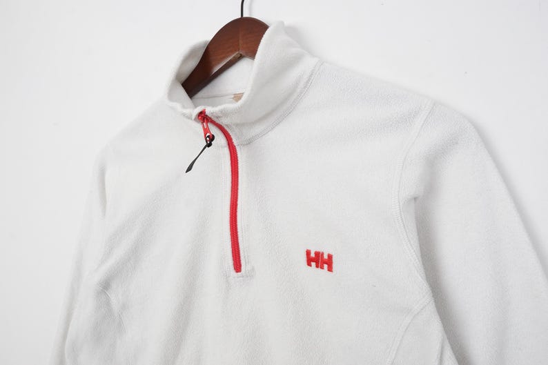 Op de afbeelding: Witte fleece trui met een hoge kraag en een rode rits. De trui heeft de geborduurde rode letters "HH" op de borst. Het kledingstuk hangt aan een houten kleerhanger.