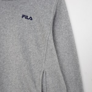 Pu&ograve; includere: Un pullover in pile grigio con collo a zip e logo Fila ricamato in blu navy. La parte superiore a maniche lunghe ha uno spacco laterale ed &egrave; realizzata in un materiale morbido e strutturato.