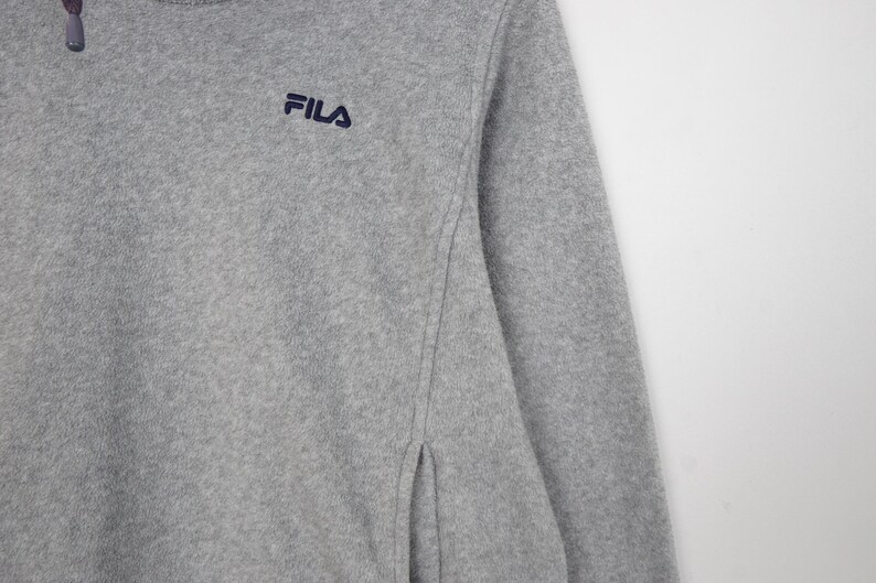 Pu&ograve; includere: Un pullover in pile grigio con collo a zip e logo Fila ricamato in blu navy. La parte superiore a maniche lunghe ha uno spacco laterale ed &egrave; realizzata in un materiale morbido e strutturato.