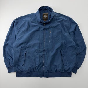 Könnte beinhalten: Eine blaue Windjacke mit durchgehendem Frontreißverschluss, Stehkragen und langen Ärmeln mit elastischen Bündchen. Die Jacke verfügt über zwei untere Vordertaschen mit Klappen sowie eine vertikale Reißverschlusstasche auf der linken Brustseite.