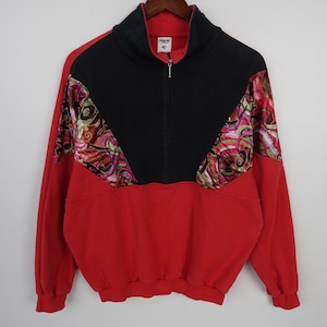 May include: A vintage red and black zip-up sweatshirt with a high collar. The upper chest and sleeves feature a colorful, patterned design. The sweatshirt is hanging on a wooden hanger.