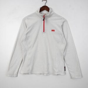 Op de afbeelding: Witte fleece trui met een rode rits en het "HH" logo. Deze top met lange mouwen heeft een opstaande kraag en is ontworpen voor buitenactiviteiten. De rits loopt over de borst.