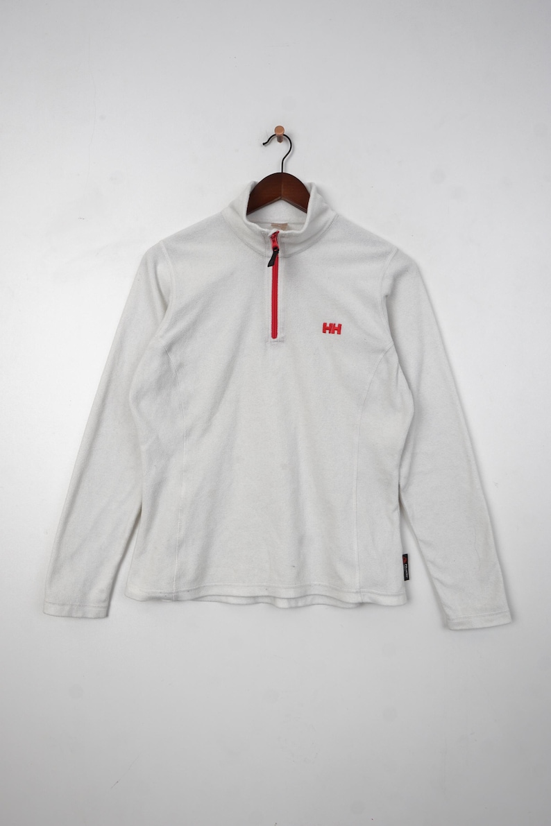 Op de afbeelding: Witte fleece trui met een rode rits en het "HH" logo. Deze top met lange mouwen heeft een opstaande kraag en is ontworpen voor buitenactiviteiten. De rits loopt over de borst.
