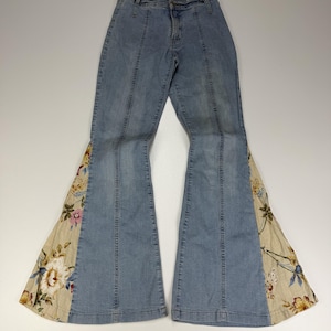 Puede incluir: Pantalones vaqueros de mezclilla azul claro con talle alto, cierre de cremallera y botón frontal. Las perneras se ensanchan en paneles acampanados de tela beige decorados con un estampado floral colorido de flores blancas, rosas y amarillas.