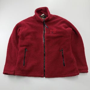 Könnte beinhalten: Eine dunkelrote Fleecejacke mit langen Ärmeln, hohem Kragen und einem durchgehenden schwarzen Reißverschluss. Die Jacke verfügt über zwei vertikale Reißverschlusstaschen auf der Vorderseite und ein kleines gesticktes Logo auf der linken Brust.