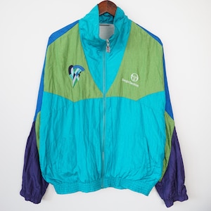 May include: A vintage teal, green, blue, and purple Sergio Tacchini track jacket. The jacket has a high collar, a full zipper, and elastic cuffs and waistband. The Sergio Tacchini logo is on the chest.