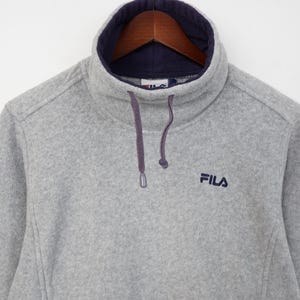 Pu&ograve; includere: Una felpa in pile grigia con collo alto foderato blu navy. Il logo Fila &egrave; ricamato in blu navy sul petto. Il colletto ha una coulisse con cordini viola.