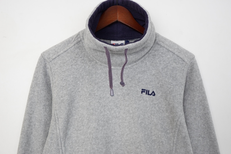 Pu&ograve; includere: Una felpa in pile grigia con collo alto foderato blu navy. Il logo Fila &egrave; ricamato in blu navy sul petto. Il colletto ha una coulisse con cordini viola.