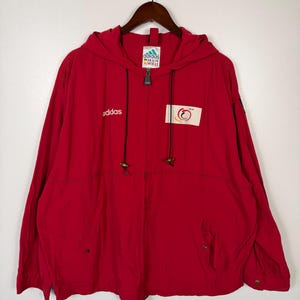 May include: A red Adidas hooded jacket with a full-length zipper and two front pockets. The jacket features the Adidas logo in white on the left chest and a patch on the right chest. The jacket is hanging on a wooden hanger.