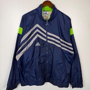 May include: Navy blue Adidas track jacket with a full zipper, gray stripes, and neon green accents. The jacket has the Adidas logo on the chest and is hanging on a wooden hanger.