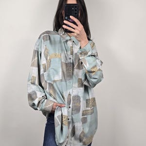 May include: A button-down shirt with a light blue and beige abstract pattern. The long-sleeved shirt has two chest pockets and is worn with blue jeans. The shirt's design includes rectangular and leaf-like shapes.