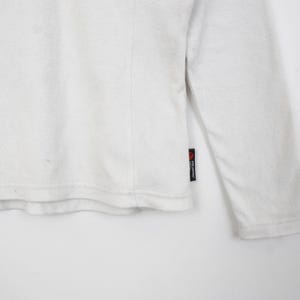 Op de afbeelding: Een witte longsleeve met een getextureerde afwerking. Het kledingstuk heeft een ronde hals en een klein zwart label met rode tekst op de mouw. De stof lijkt zacht te zijn, mogelijk fleece.