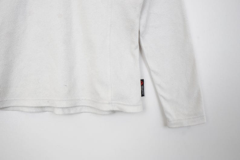 Op de afbeelding: Een witte longsleeve met een getextureerde afwerking. Het kledingstuk heeft een ronde hals en een klein zwart label met rode tekst op de mouw. De stof lijkt zacht te zijn, mogelijk fleece.